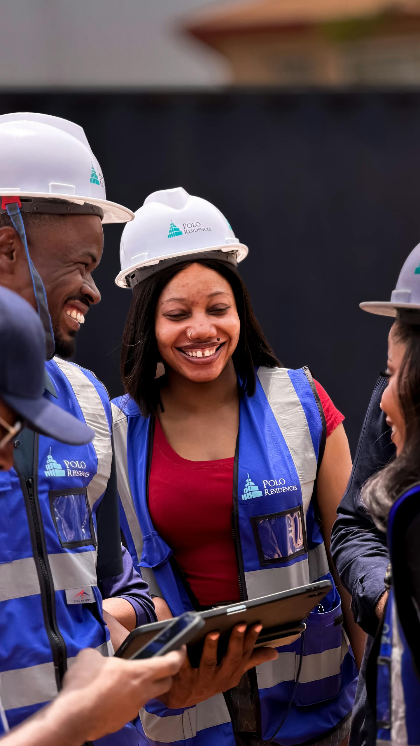 A group of construction workers collaborating and smiling outdoors, wearing safety gear and using a tablet.